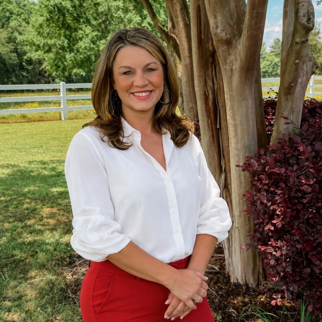 A woman in white blouse and red pants stands outside near a tree, smiling at the camera.