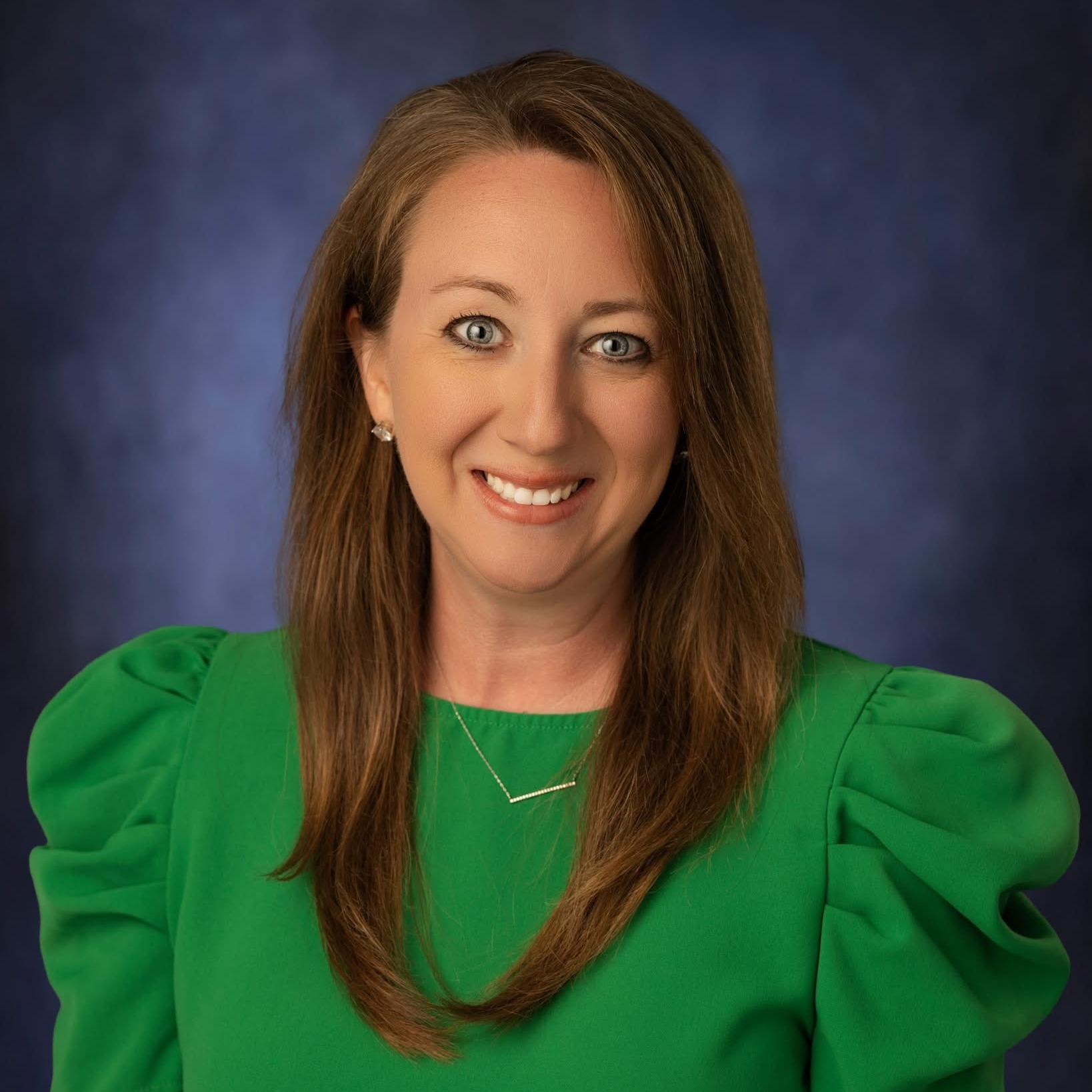 Woman with brown hair, wearing a green blouse with puffed sleeves, smiling, against a blue background.