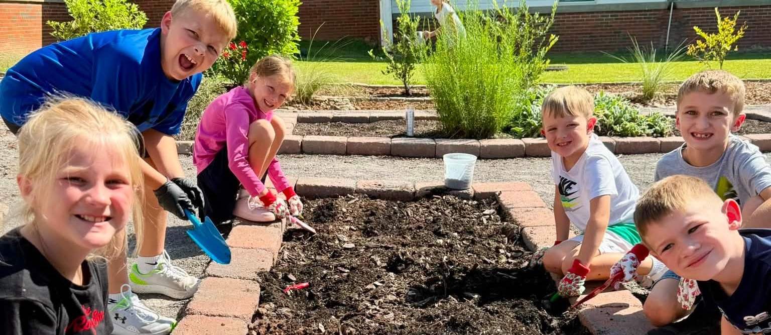 Group of children planting in a garden. Some children wear gloves. A building is in the background.
