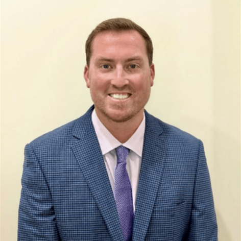 Man in a blue suit and purple tie, smiling with hands in front of him. Background is plain white.