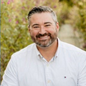 Man with short gray hair and beard smiles against a blurred background of pink flowers.
