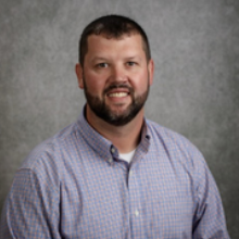 A man with a beard and short hair smiles in front of a gray background. He wears a blue checkered shirt.