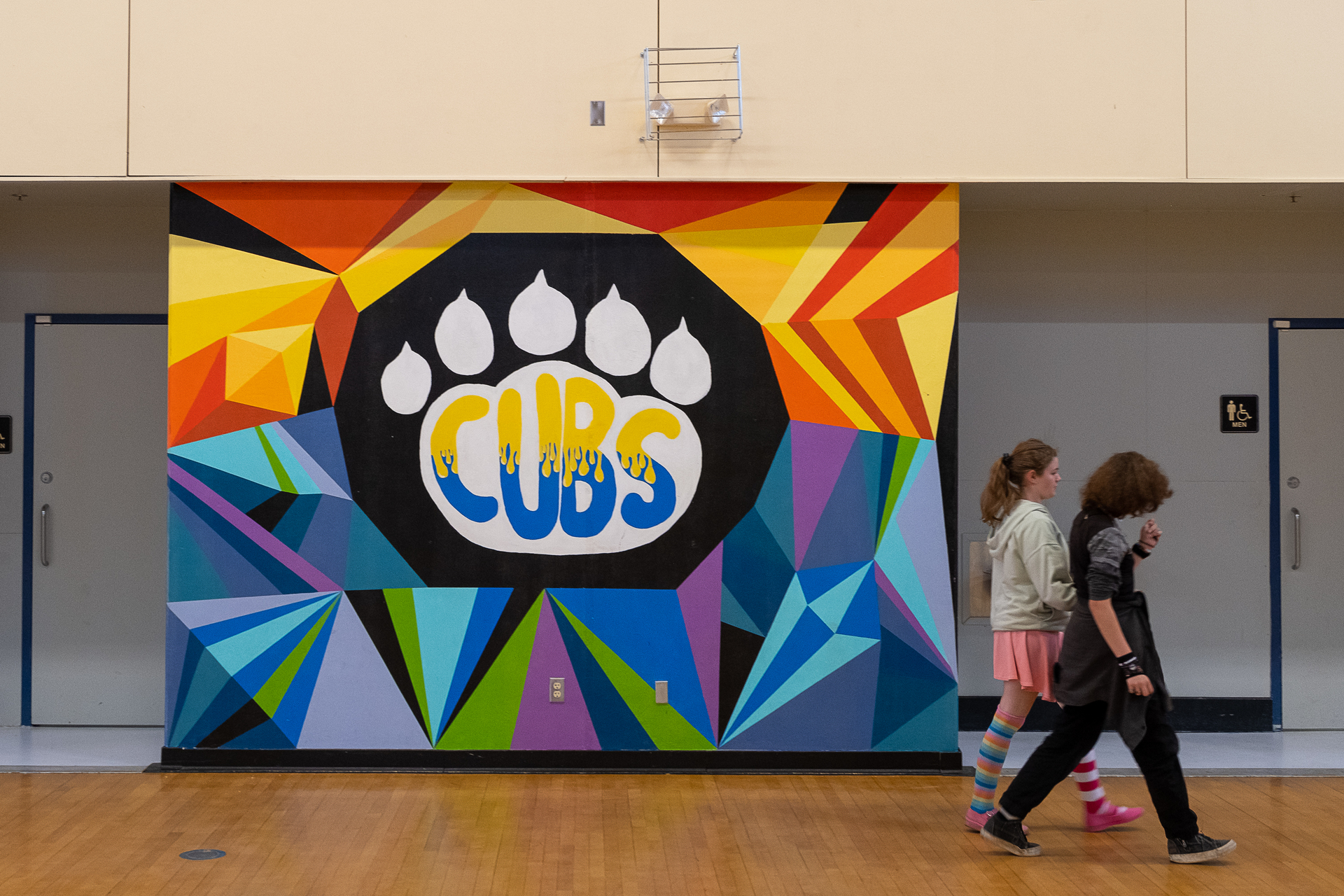 Colorful School Mural: Two students walk past a vividly painted mural with geometric shapes and the word "CUBS" in a school gymnasium, embodying school spirit and artistic flair.