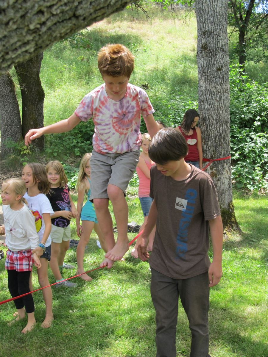 A young boy with red hair in a tie-dye shirt is carefully walking on a slackline, holding the hand of a boy in a grey shirt who is assisting him. They are surrounded by a group of cheering children in a grassy area with trees in the background.