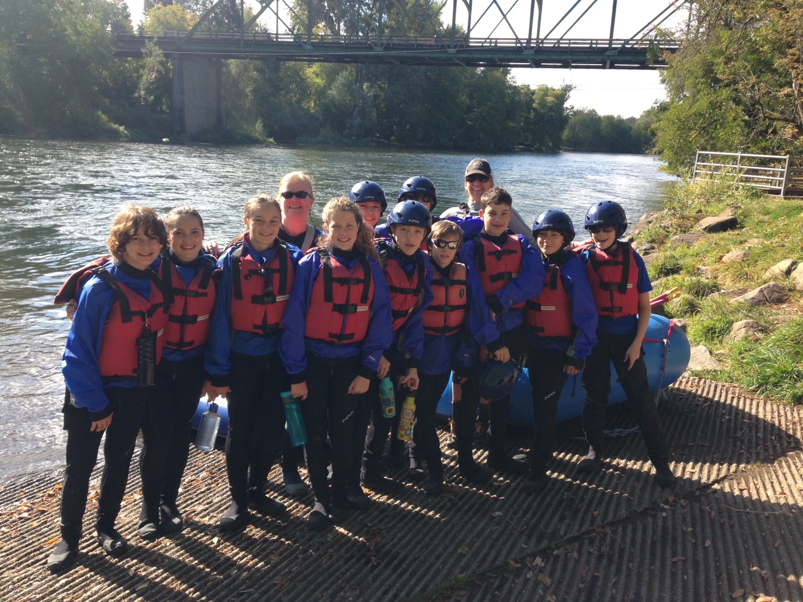 A group of students wearing blue life jackets and helmets poses for a photo by a river. A bridge and trees are in the background, suggesting preparation for a water activity, possibly rafting.