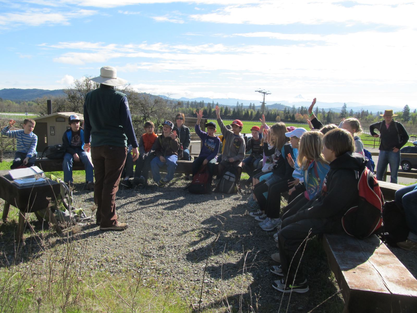  group of students sitting on benches outdoors attentively listen to an instructor wearing a hat. They are raising their hands, ready to answer or ask questions. The background shows a clear sky and a distant mountain range.