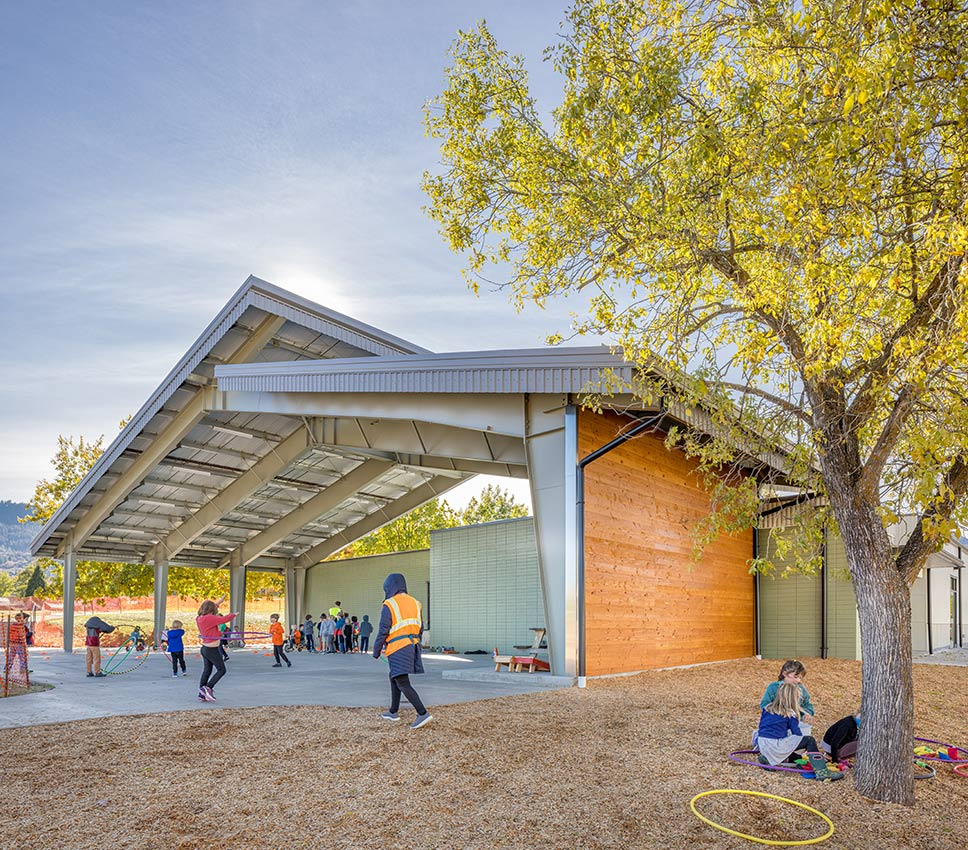 Outdoor play area at a school with children engaged in various activities. A large, modern pavilion with a sloped metal roof provides shelter. The pavilion is supported by steel columns and open on all sides, with one wall featuring a warm wooden finish. A tree with yellowing leaves suggests an autumn season. Children in the foreground are playing with hula hoops, while others in the background are gathered in a group activity. The ground is covered in wood chips for safety, and the sky is clear, indicating a sunny day.