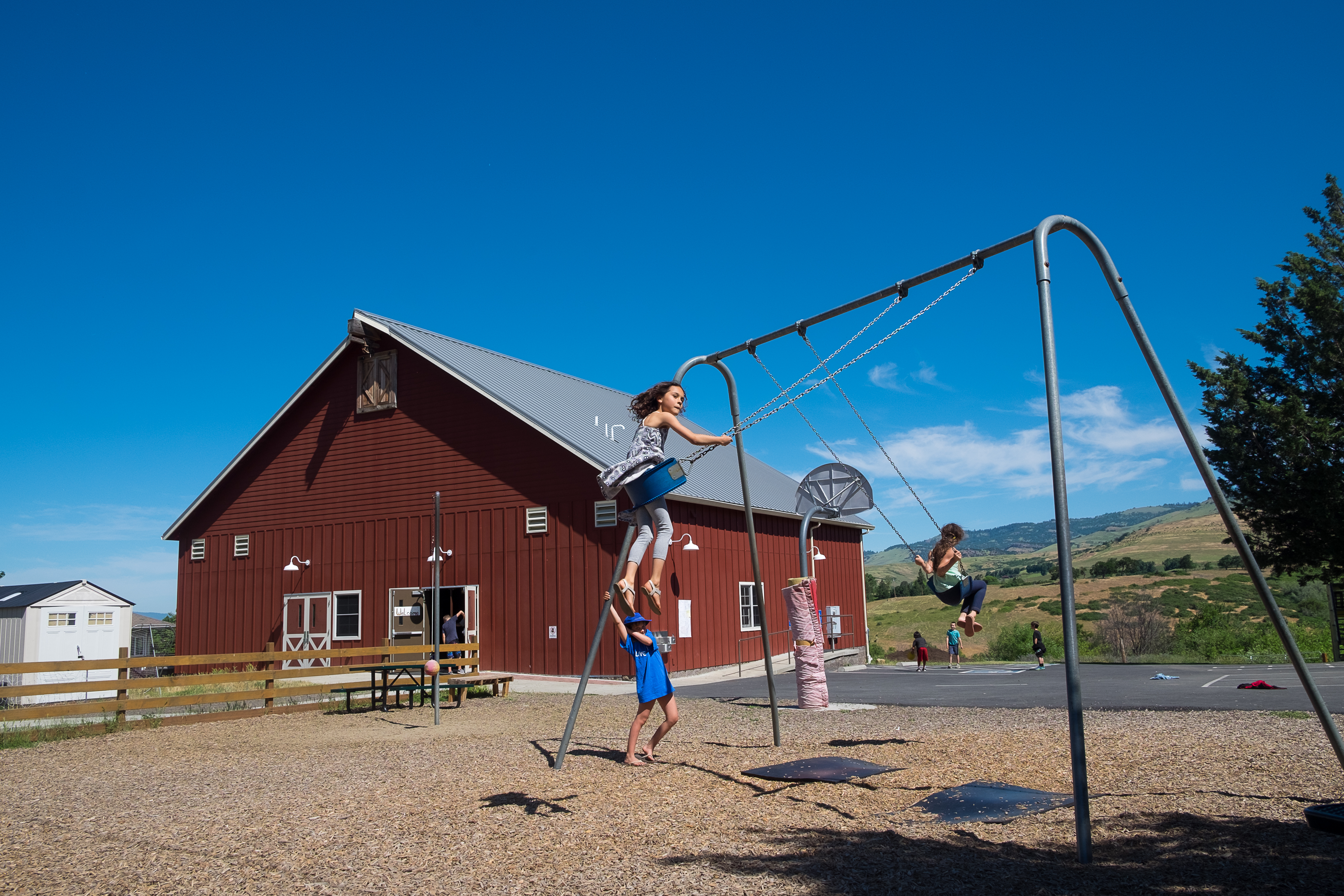 Children playing on swings in a sunny playground with a clear blue sky. A large red barn and other outbuildings are in the background, creating a rural setting.