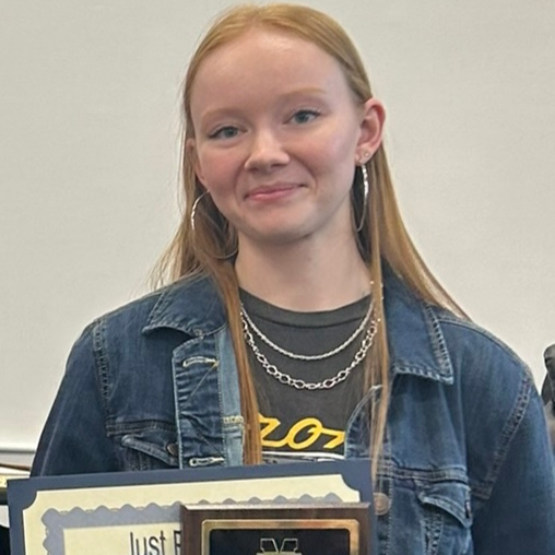 Young woman with long red hair, wearing a denim jacket holding an award.