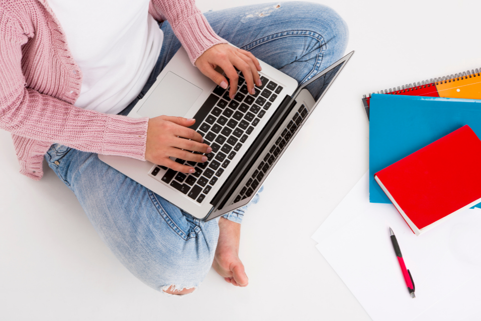 A laptop on a girls lap with books and a pen in front of her.