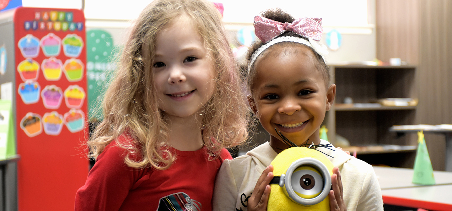 Two children stand together in a classroom, looking at the camera