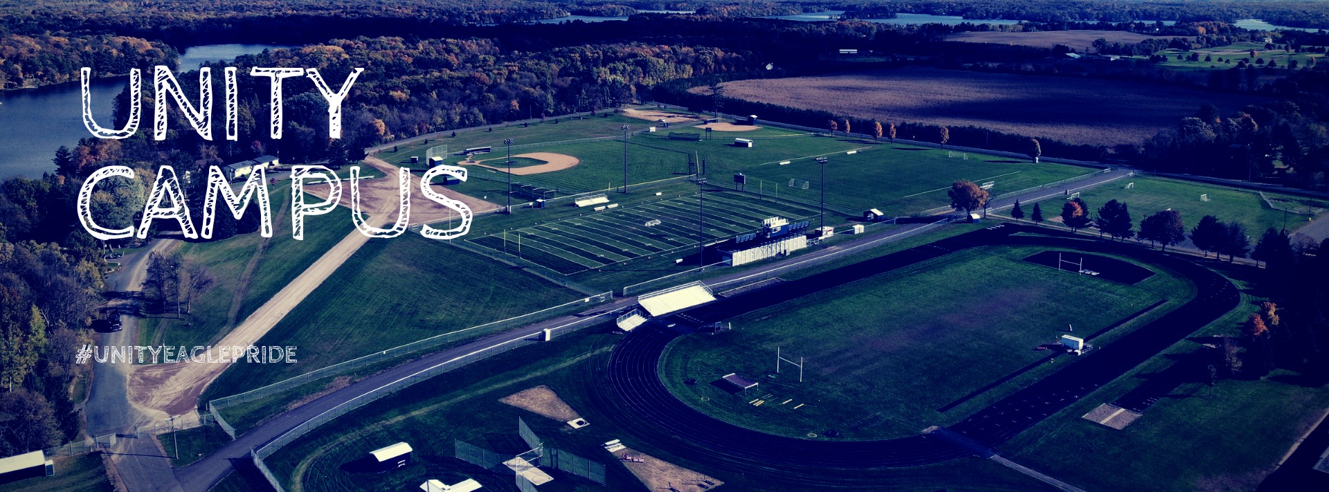 Aerial view of the Unity School District campus showing the athletic tracks, green fields, and surrounding forest with the text "Unity Campus" overlaid