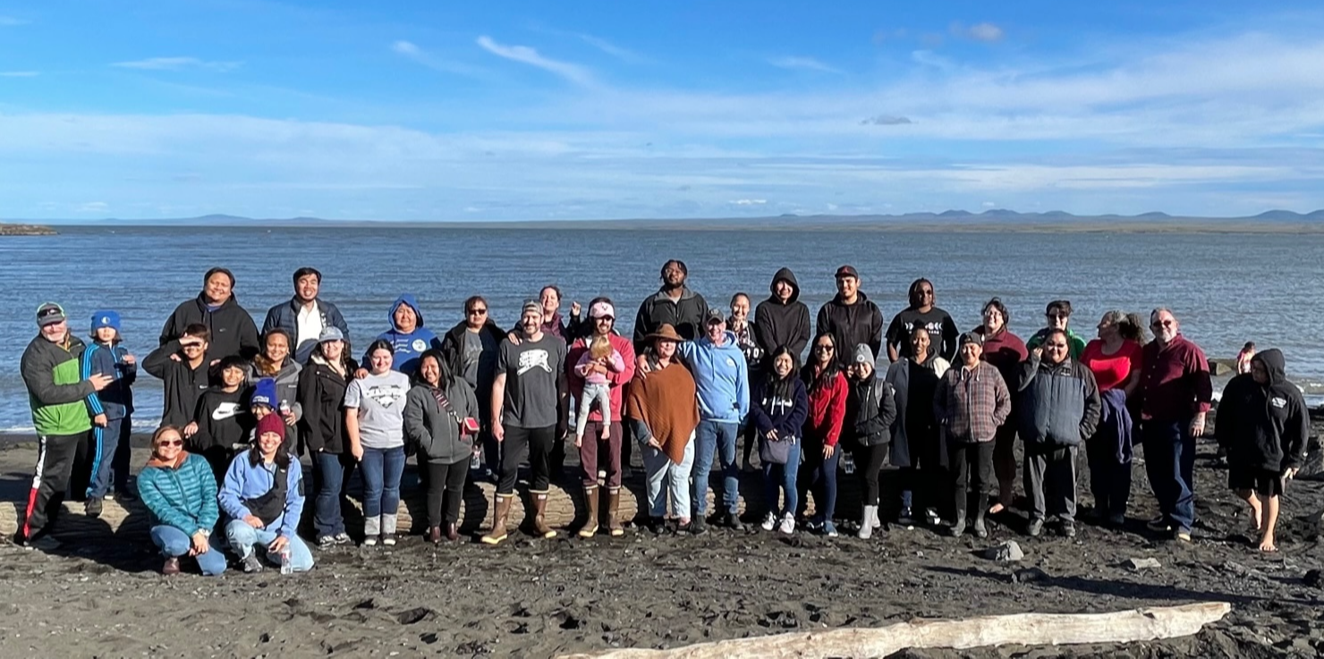 Staff from Stebbins and St Michael pose under the blue sky, on the sand, in front of the ocean with mountains on the distant horizon.