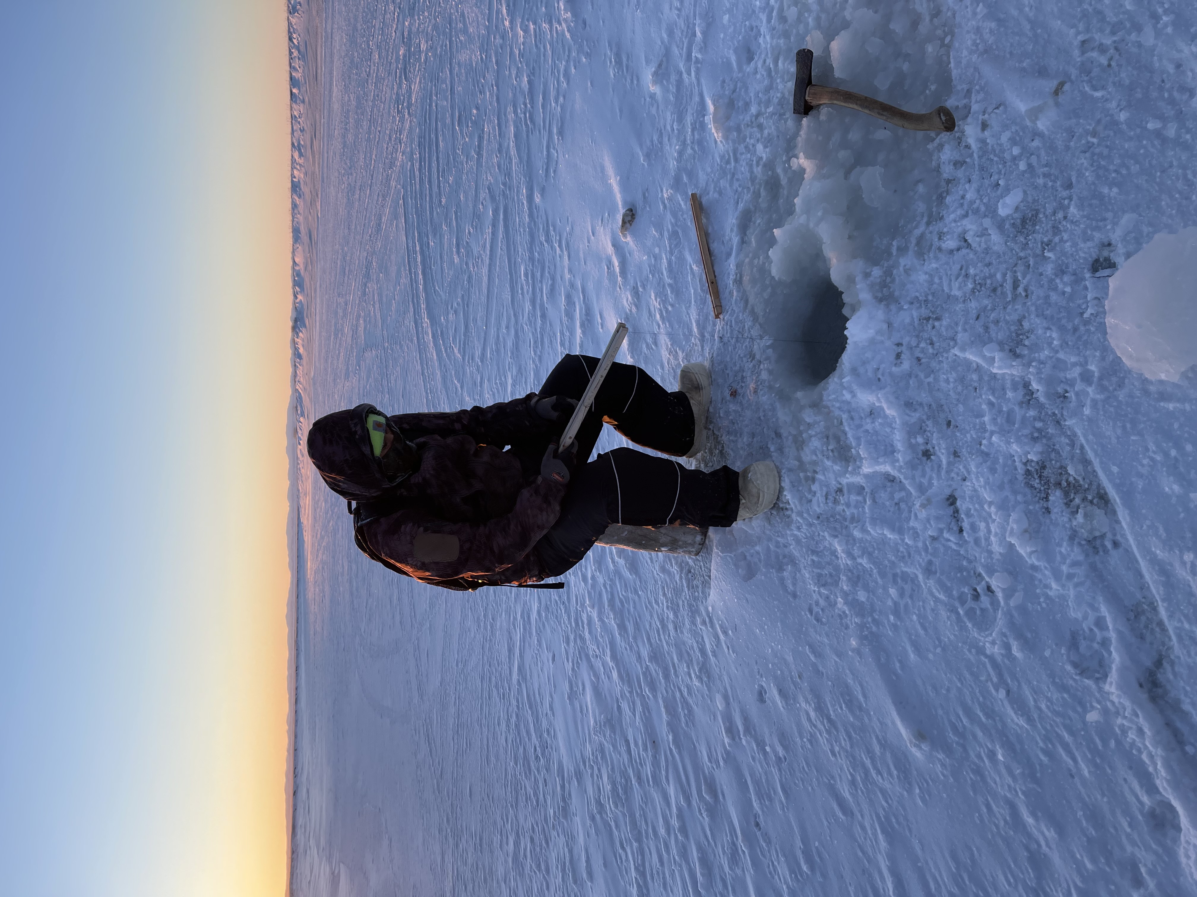 A teen sits on a bucket over an ice hole holding a fishing line in the dusk just after sunset.