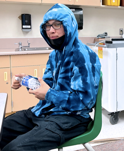 A seated student in a blue hoody hydrates with bottled water.