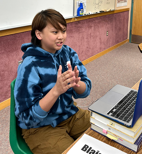 A student seated before a laptop gestures with his hands during a public speaking competition via Zoom.