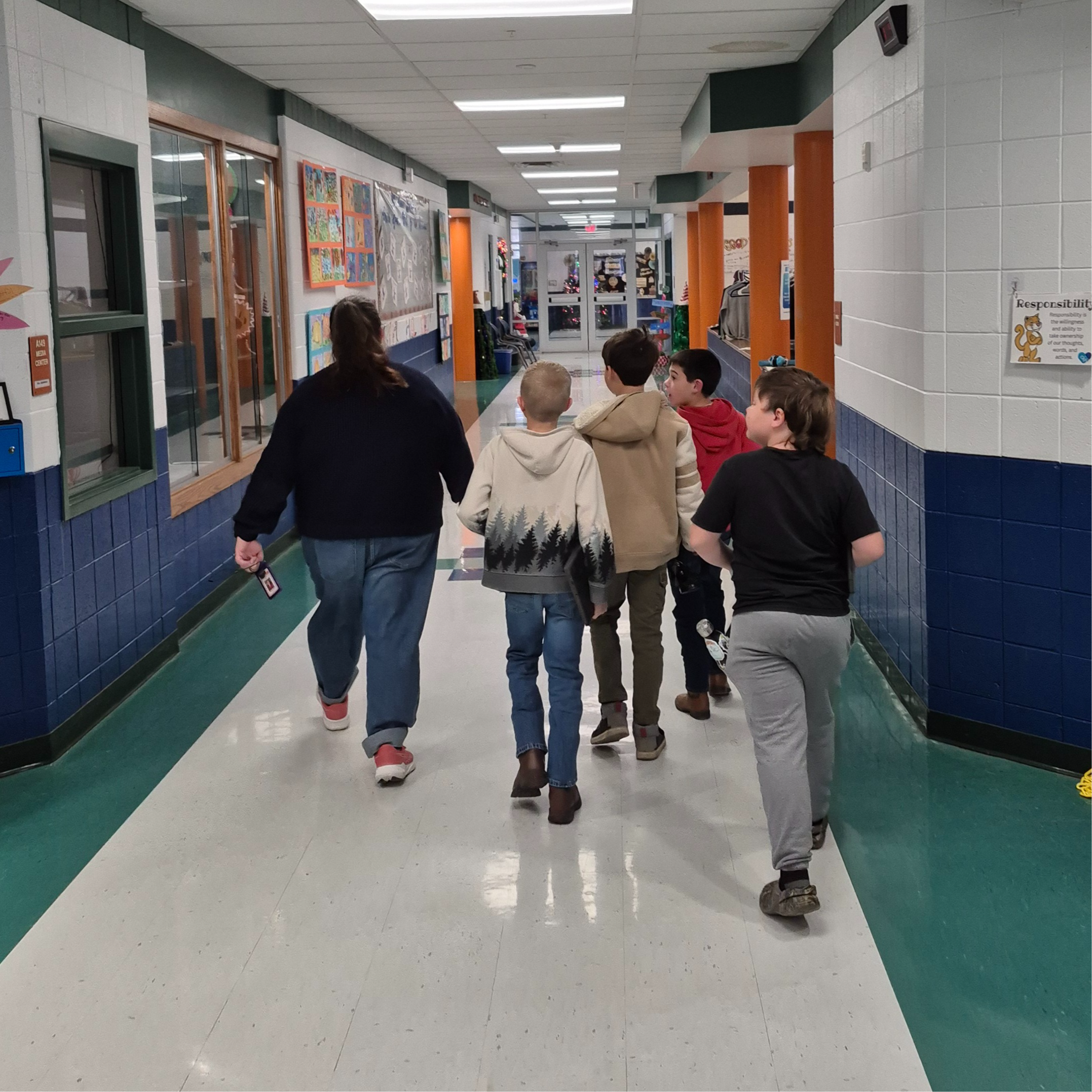 A teacher walks down a school hallway with a small group of students, engaging with them as they return to class. Colorful bulletin boards and classroom doors line the corridor.
