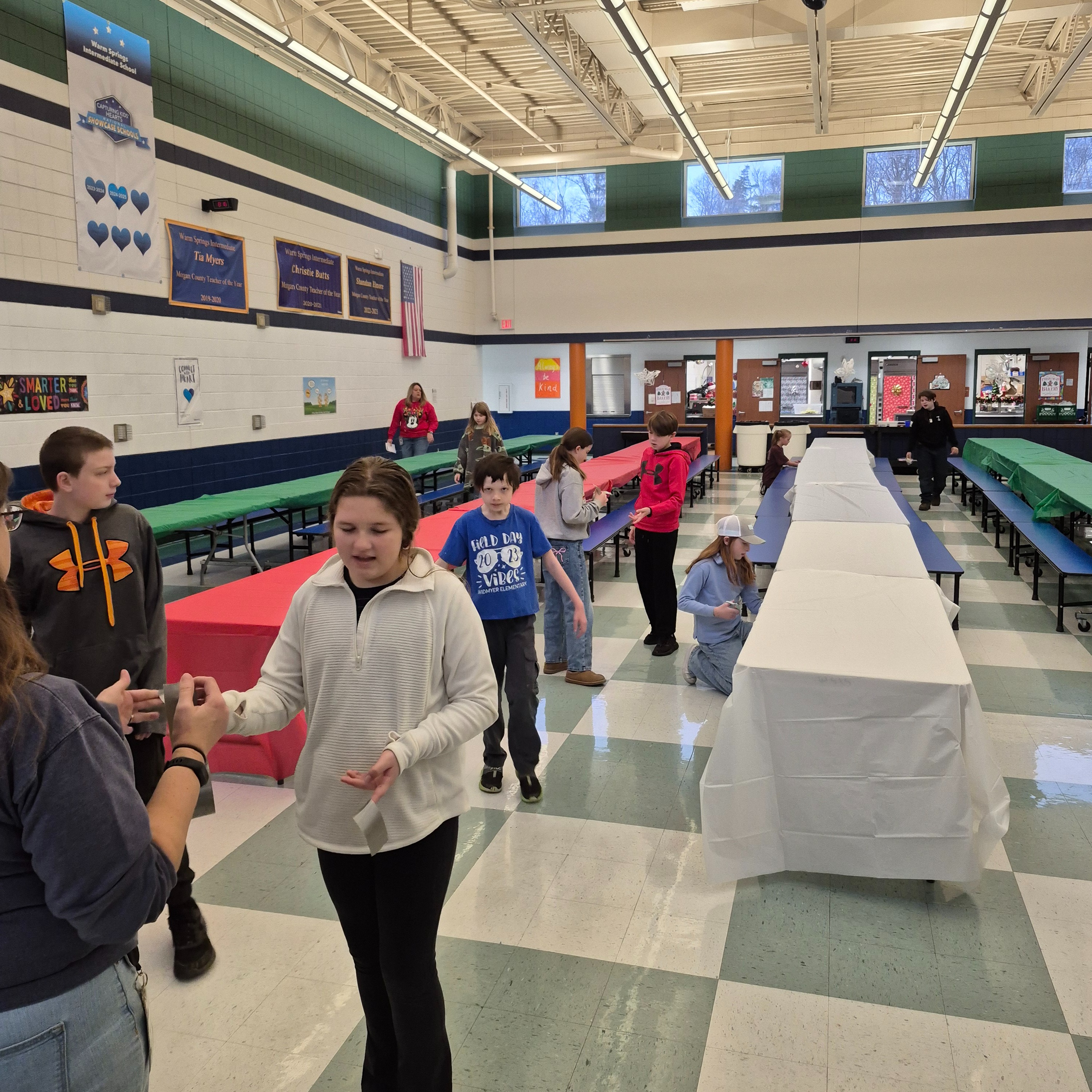 Students work together in the school cafeteria, setting up tables with tablecloths and preparing the space for an upcoming special event.
