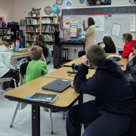 Students sit at tables attentively as a teacher leads a classroom lesson, with student participation and visual displays supporting engagement and discussion.