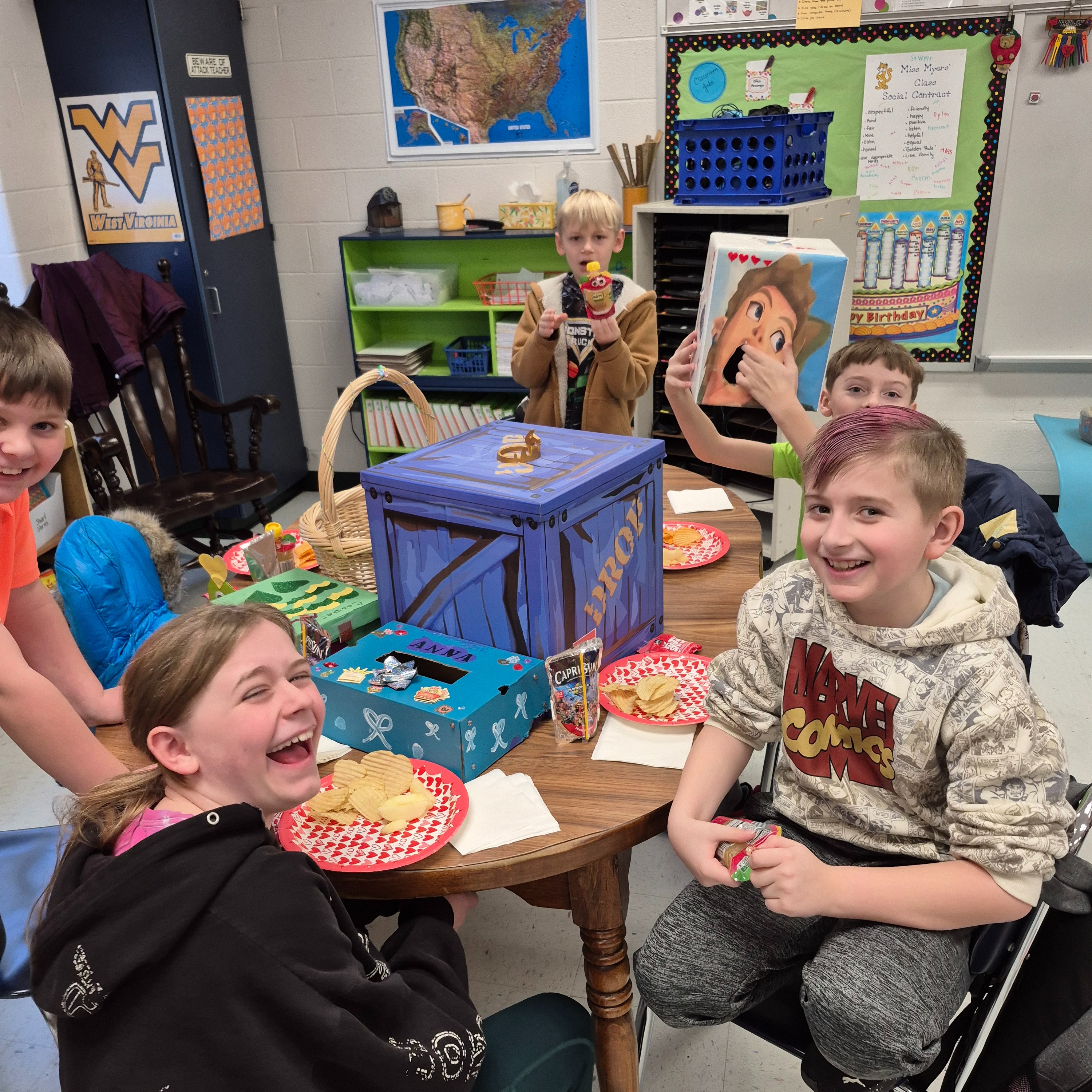 Students smiling and laughing around a classroom table during a celebration, with snacks, decorated boxes, and Valentine-themed items displayed.