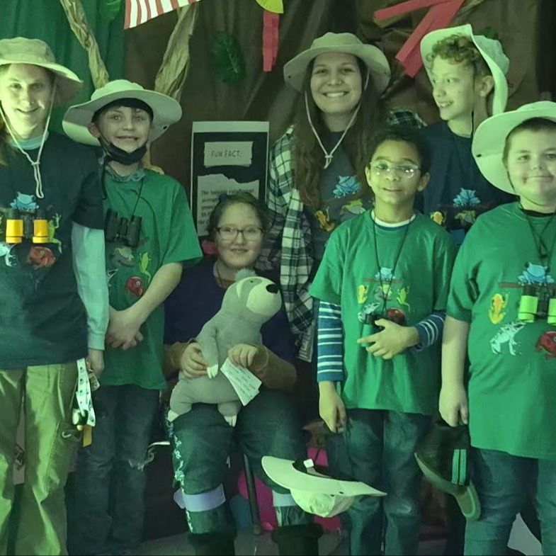 Six students and their teacher pose in safari hats and green shirts before a jungle-themed display. One student holds a stuffed sloth as the group smiles proudly.