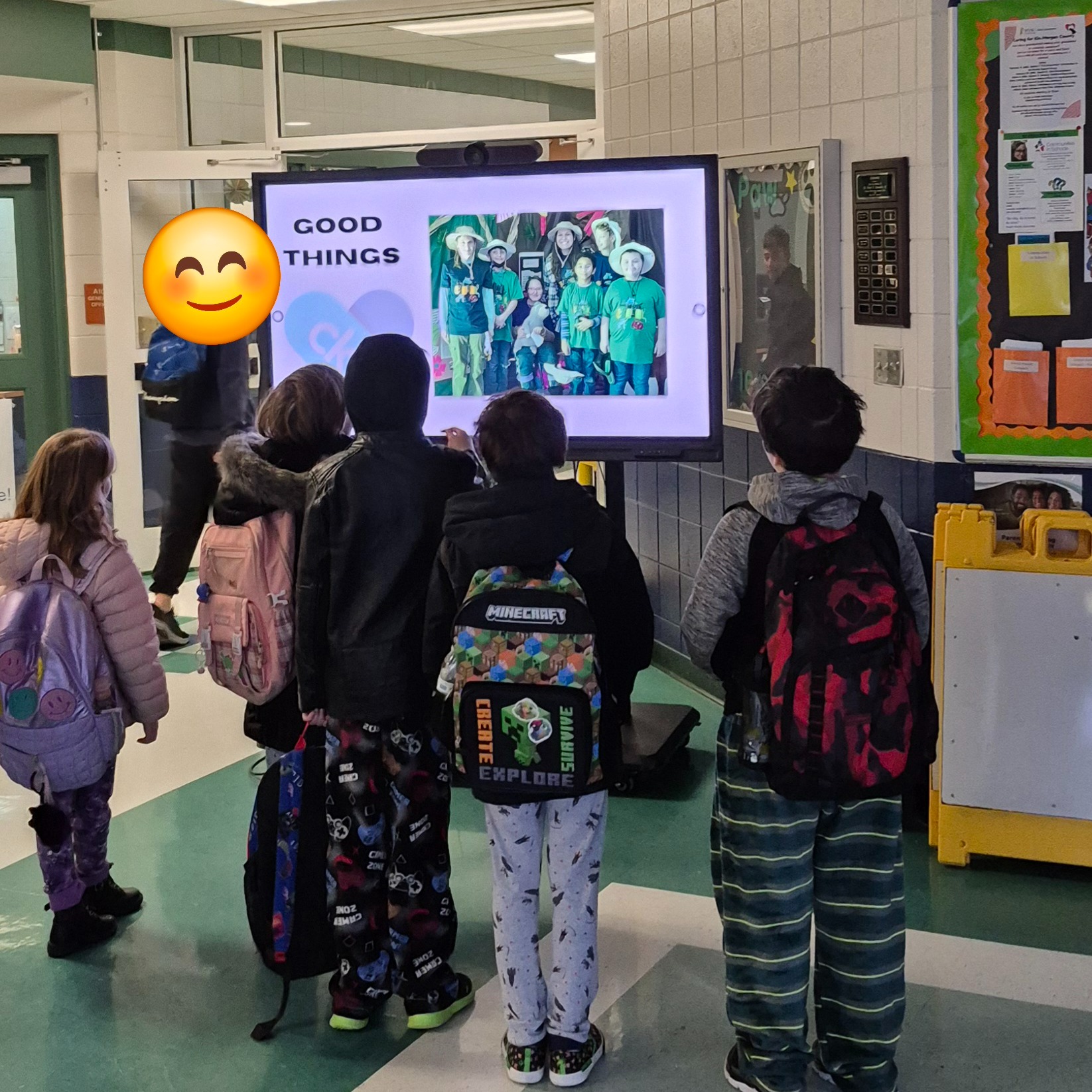 Students wearing backpacks gather in a school hallway watching a “Good Things” slideshow displayed on a screen, celebrating positive moments and student achievements at Warm Springs Intermediate School.