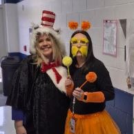 Two staff members dressed in playful costumes, including a Cat in the Hat outfit and colorful accessories, smile and pose together in a school hallway during Read Across America Week.