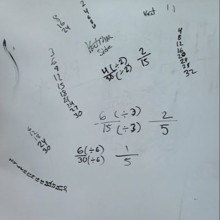 Whiteboard showing math work on equivalent fractions, including dividing numerators and denominators, factor lists, and examples like 2/15, 2/5, and 1/5