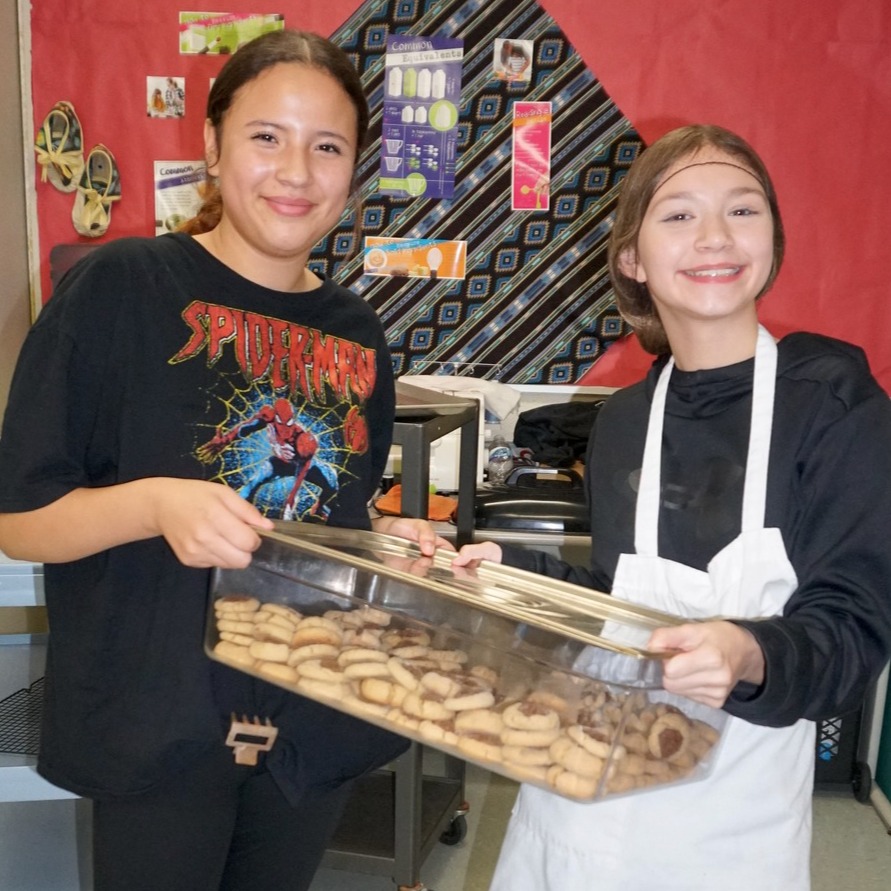 two students wearing aprons smile while holding a tray of cookies