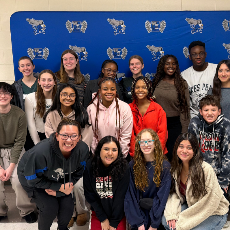 A group of young people stand in front of a blue backdrop with a school logo.
