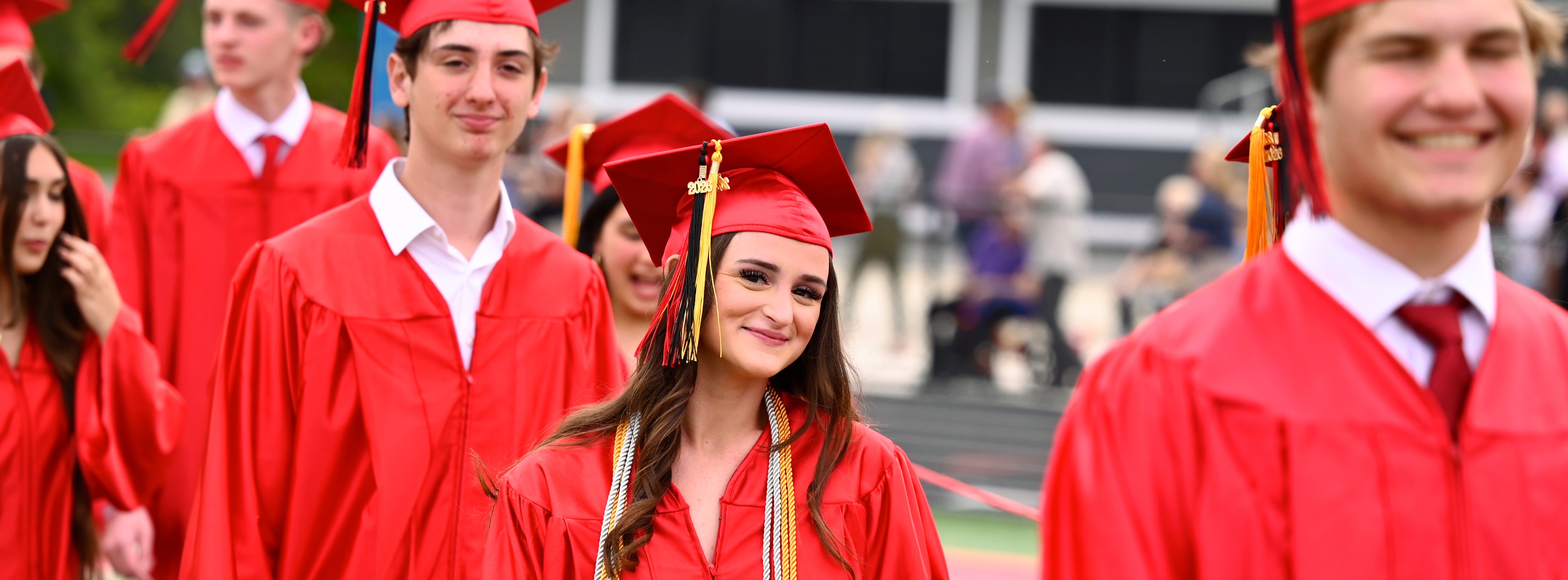 Graduates in red caps and gowns smile and walk in a line on a sports field.