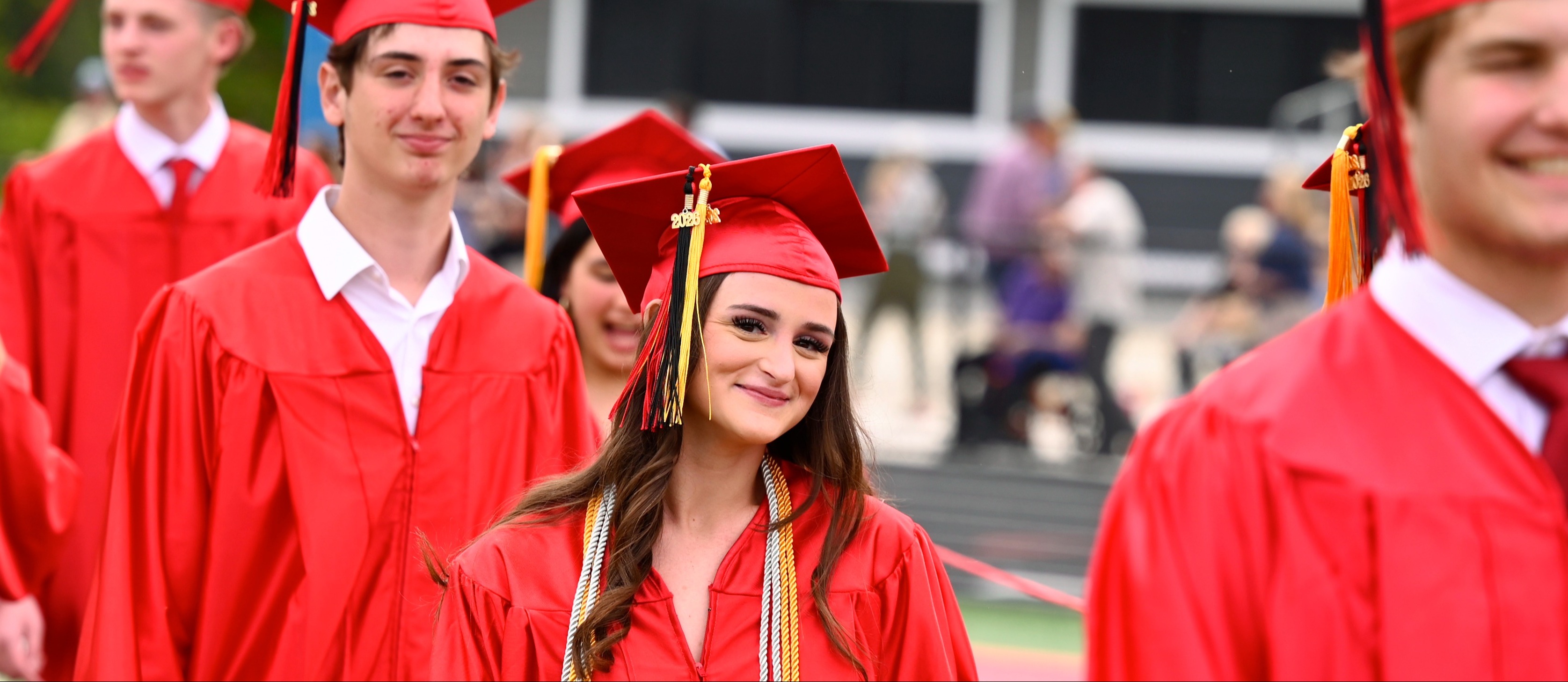 Graduates in red caps and gowns smile and walk in a line on a sports field.