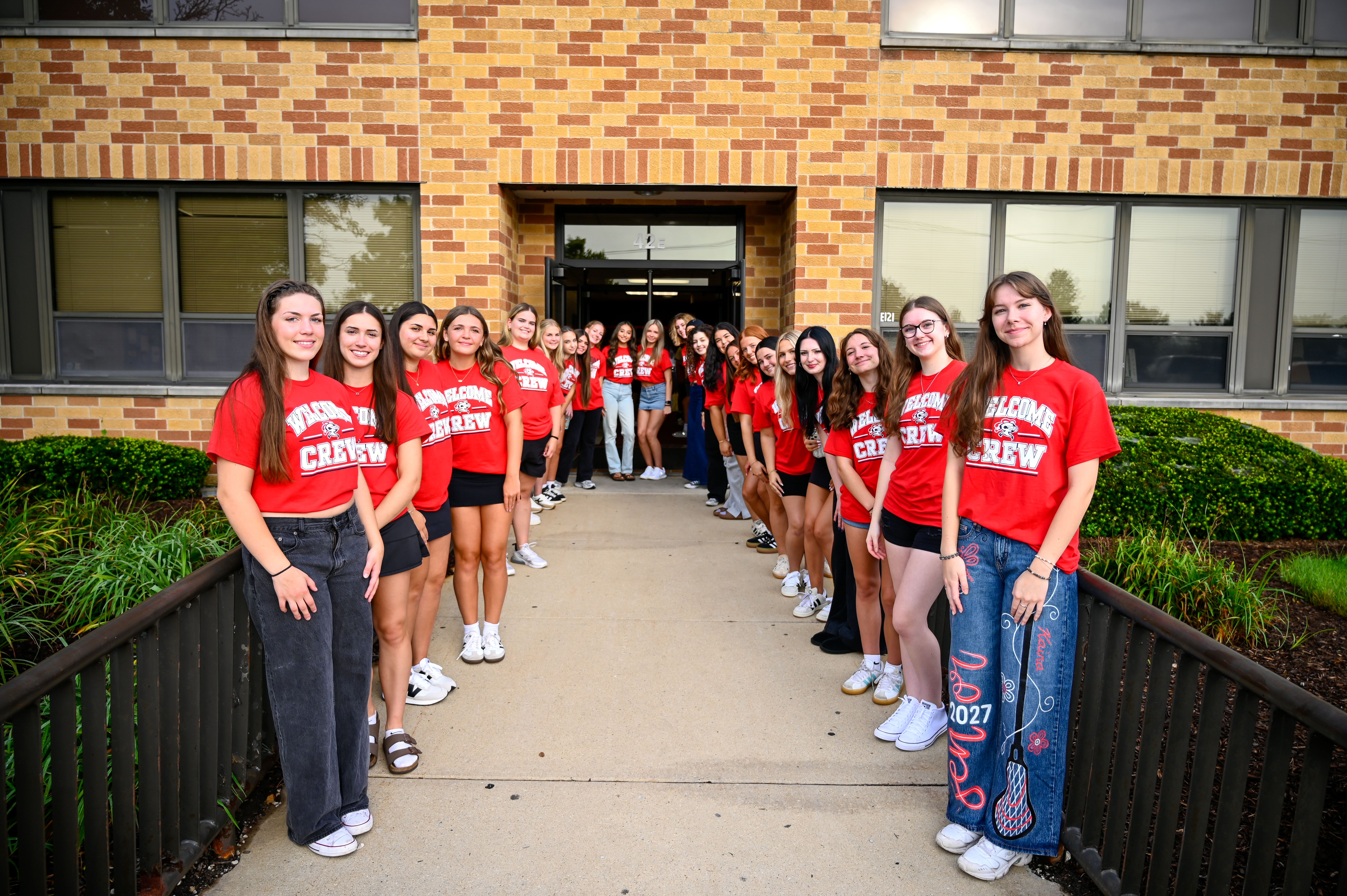 A line of young women in red shirts and jeans stand in front of a brick building.