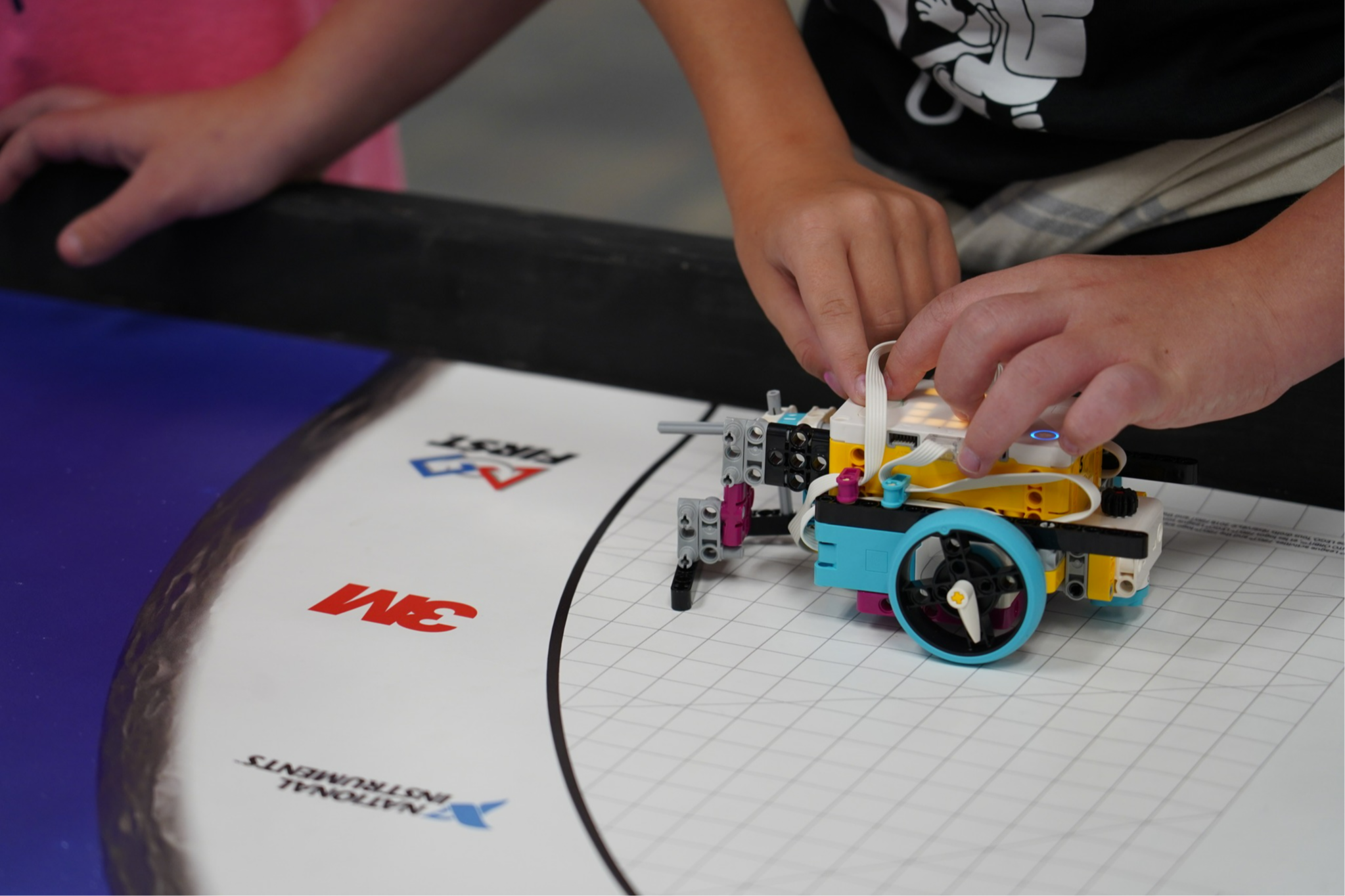 Hands work on a robot on a square mat with a track. Logos are visible on the mat.