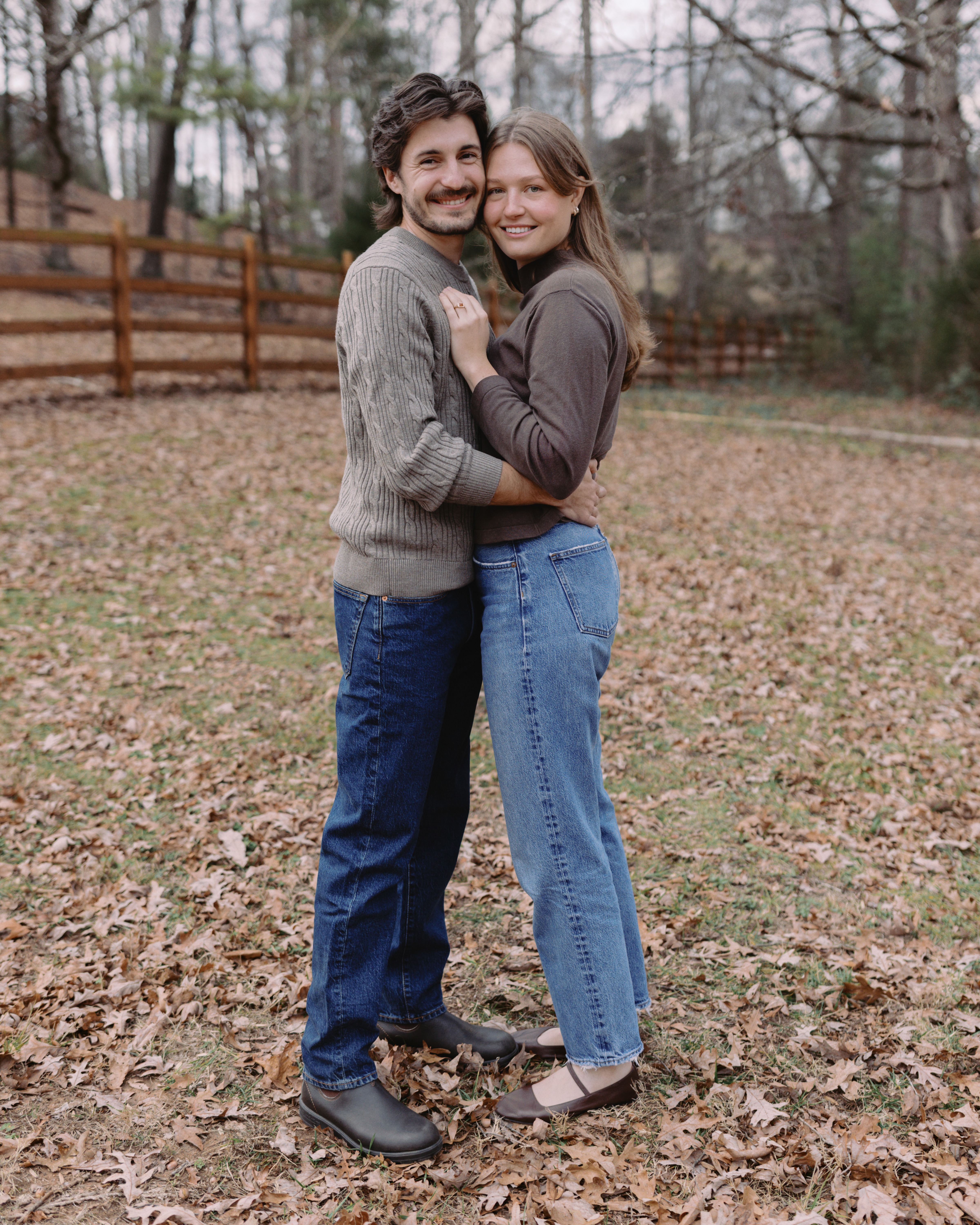 A couple stands close together in an open field with dried leaves, wearing jeans and sweaters. Trees and a fence in the background.