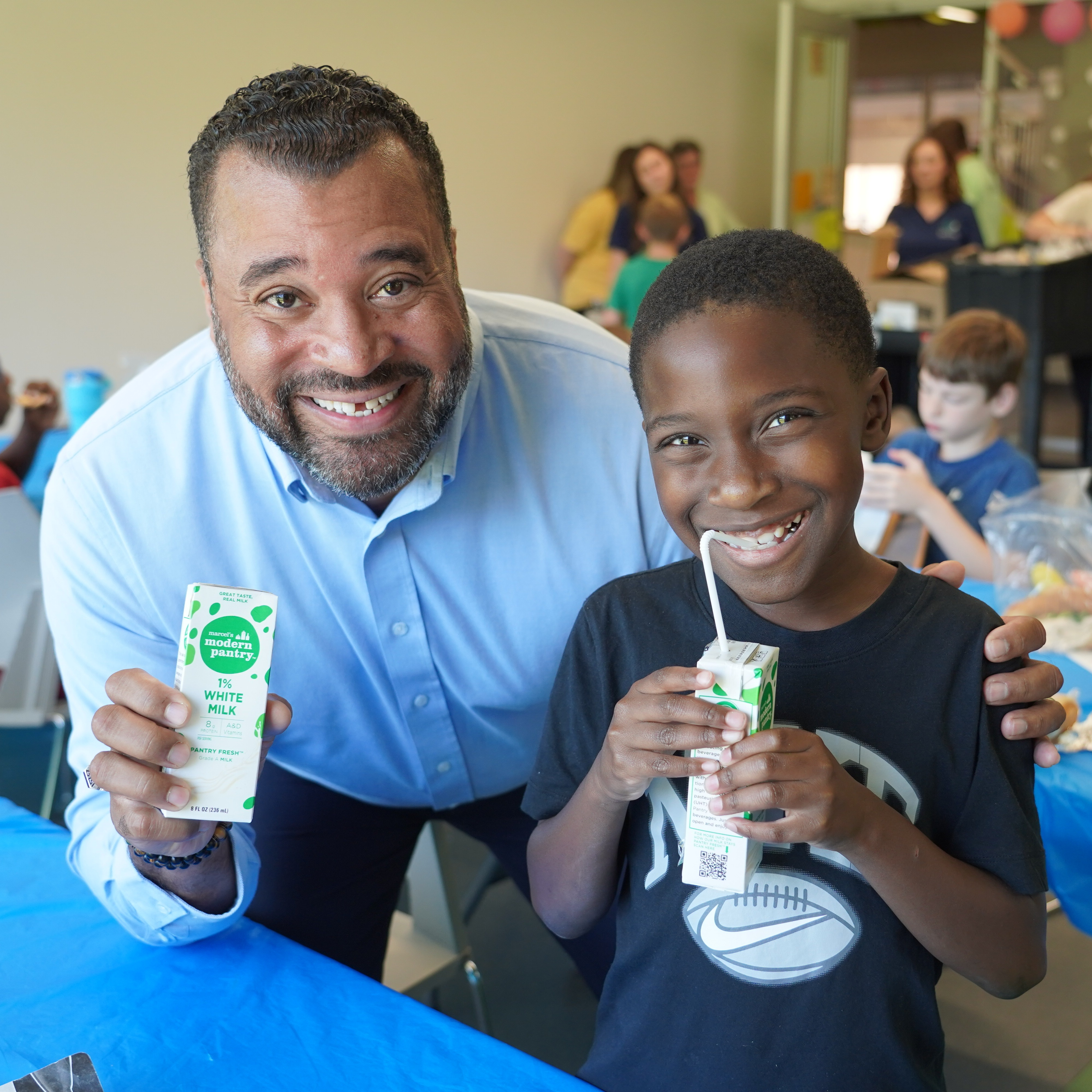 A smiling man and boy pose with cartons of milk in a room with tables and chairs.