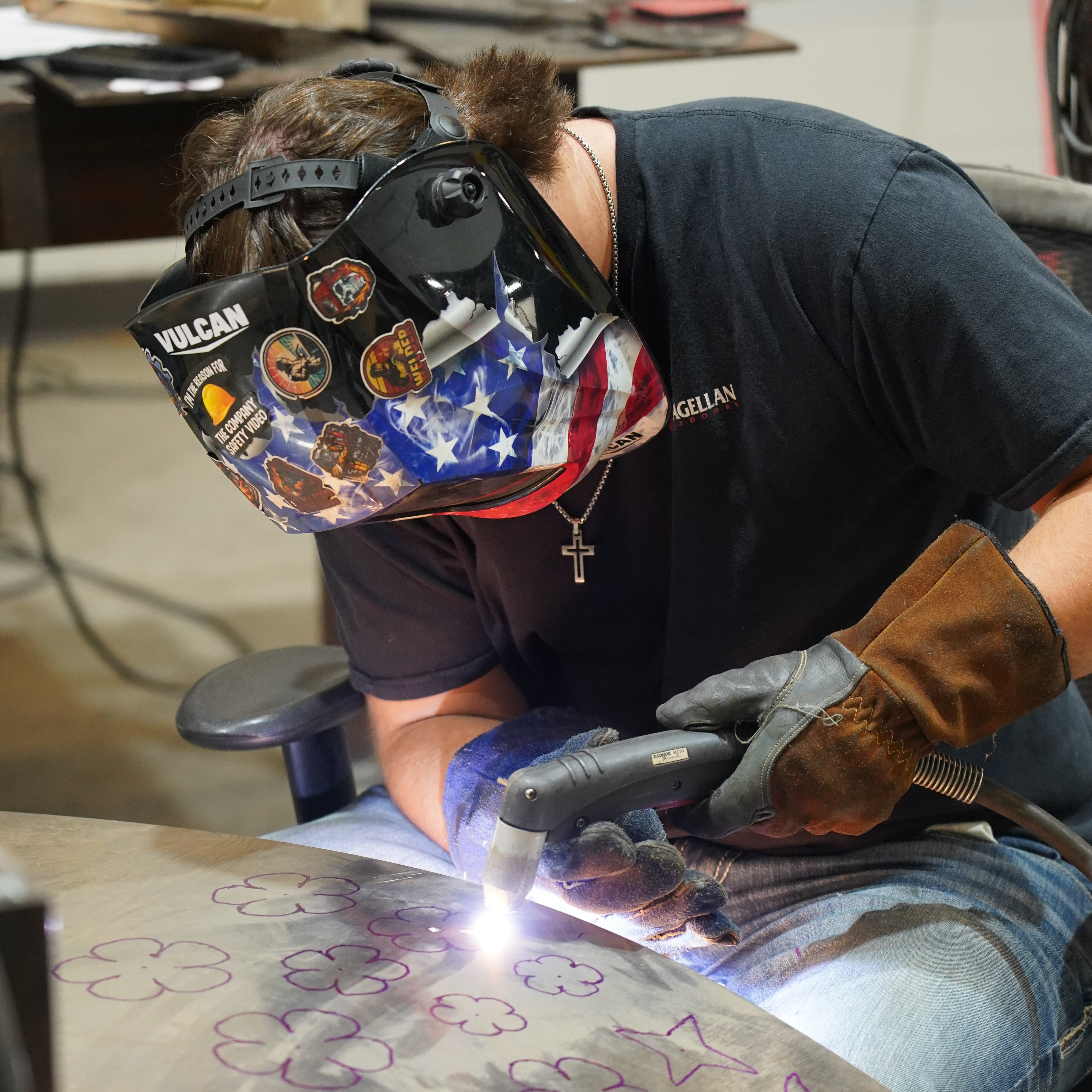 Person wearing safety gear and welding a metal piece with mosaic pattern on a table.