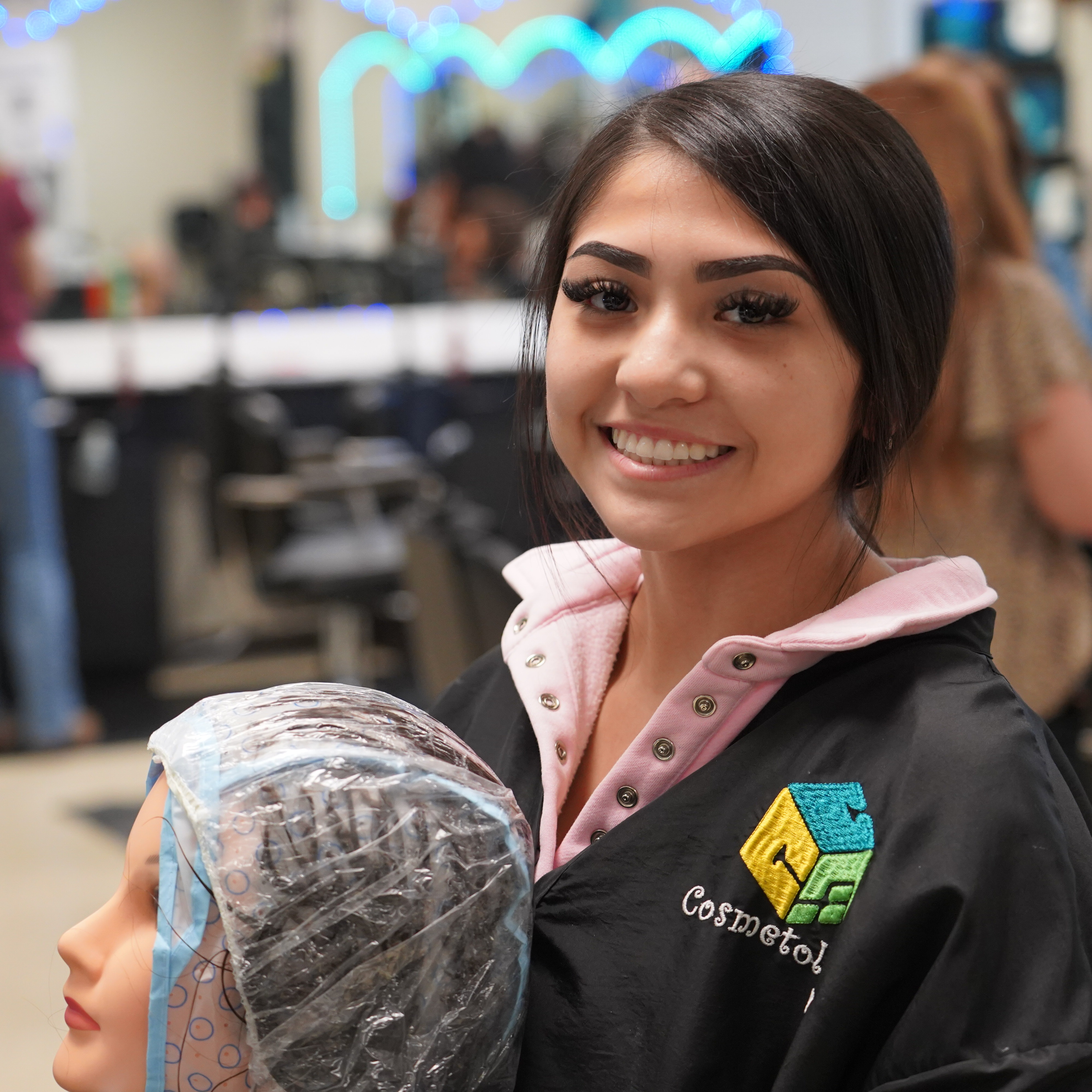 A woman holds a hair model covered with plastic wrap. Behind her, in a salon, several people stand and work.