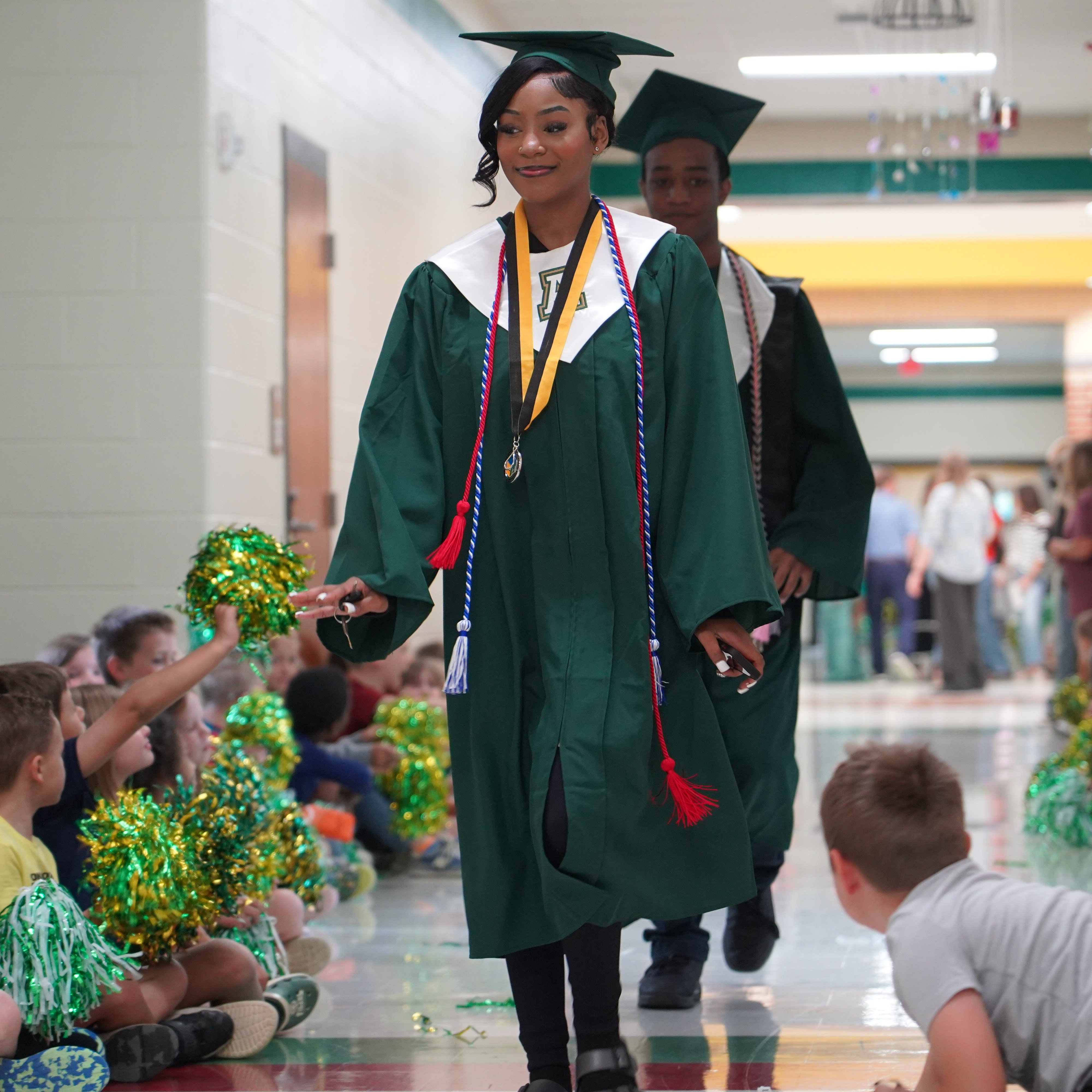 A hallway with students seated on the floor. A graduate in a green gown walks past them.
