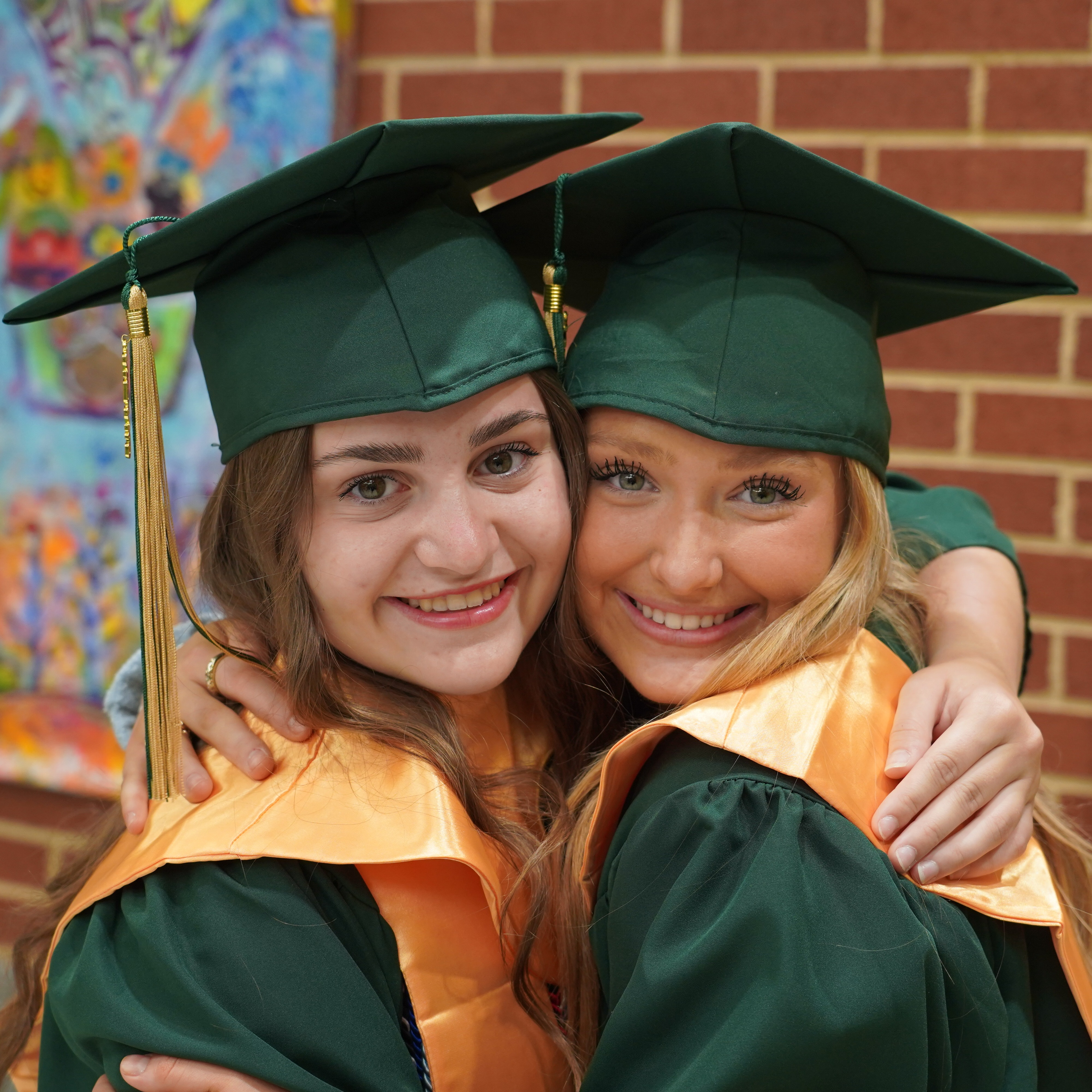 Two women in caps and gowns smiling at each other in front of a brick wall.