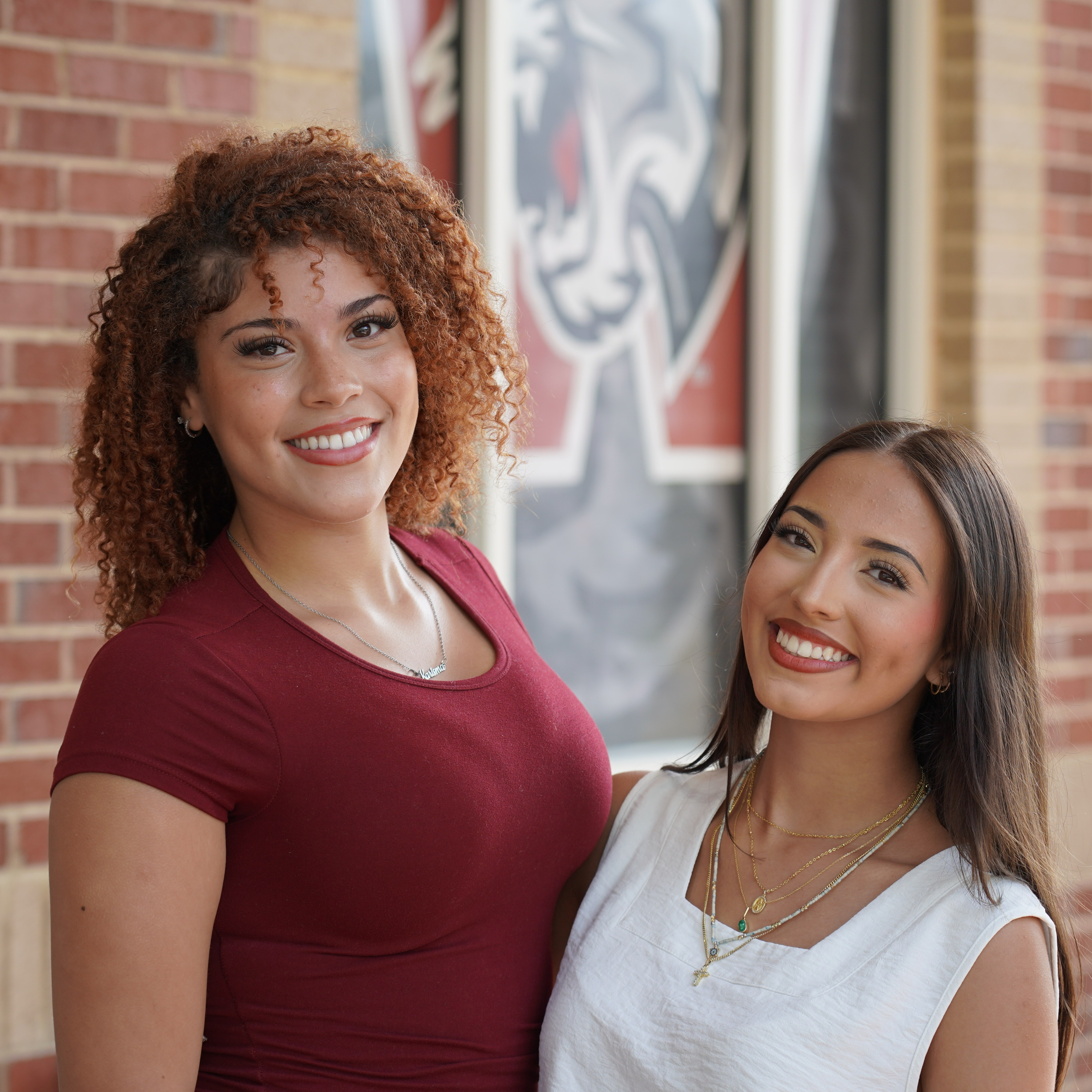 Two women smile in front of a brick wall with a university logo, one wears a red shirt, the other a white top.