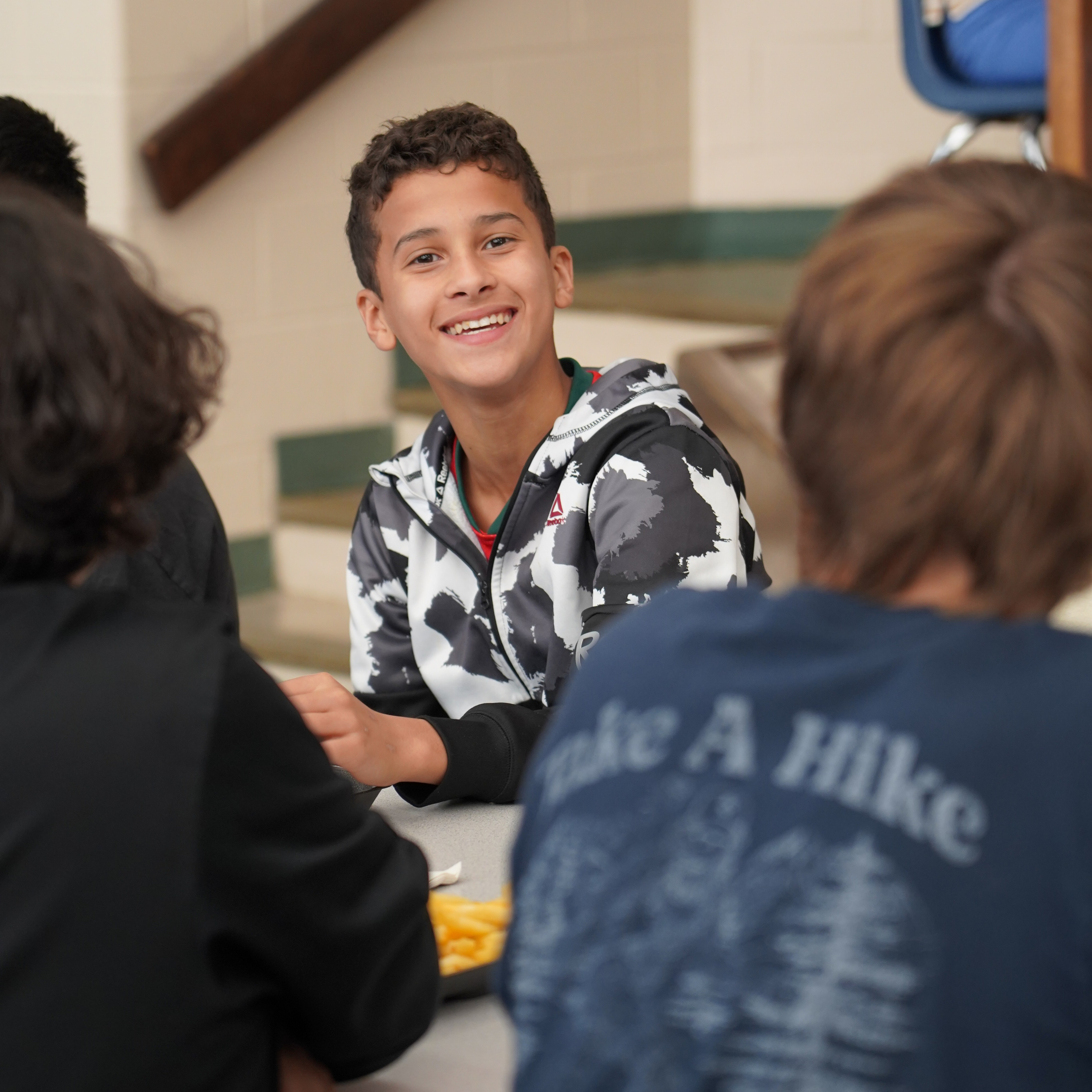 Group of students seated in a cafeteria. A boy smiles at others, with two of them wearing black shirts.