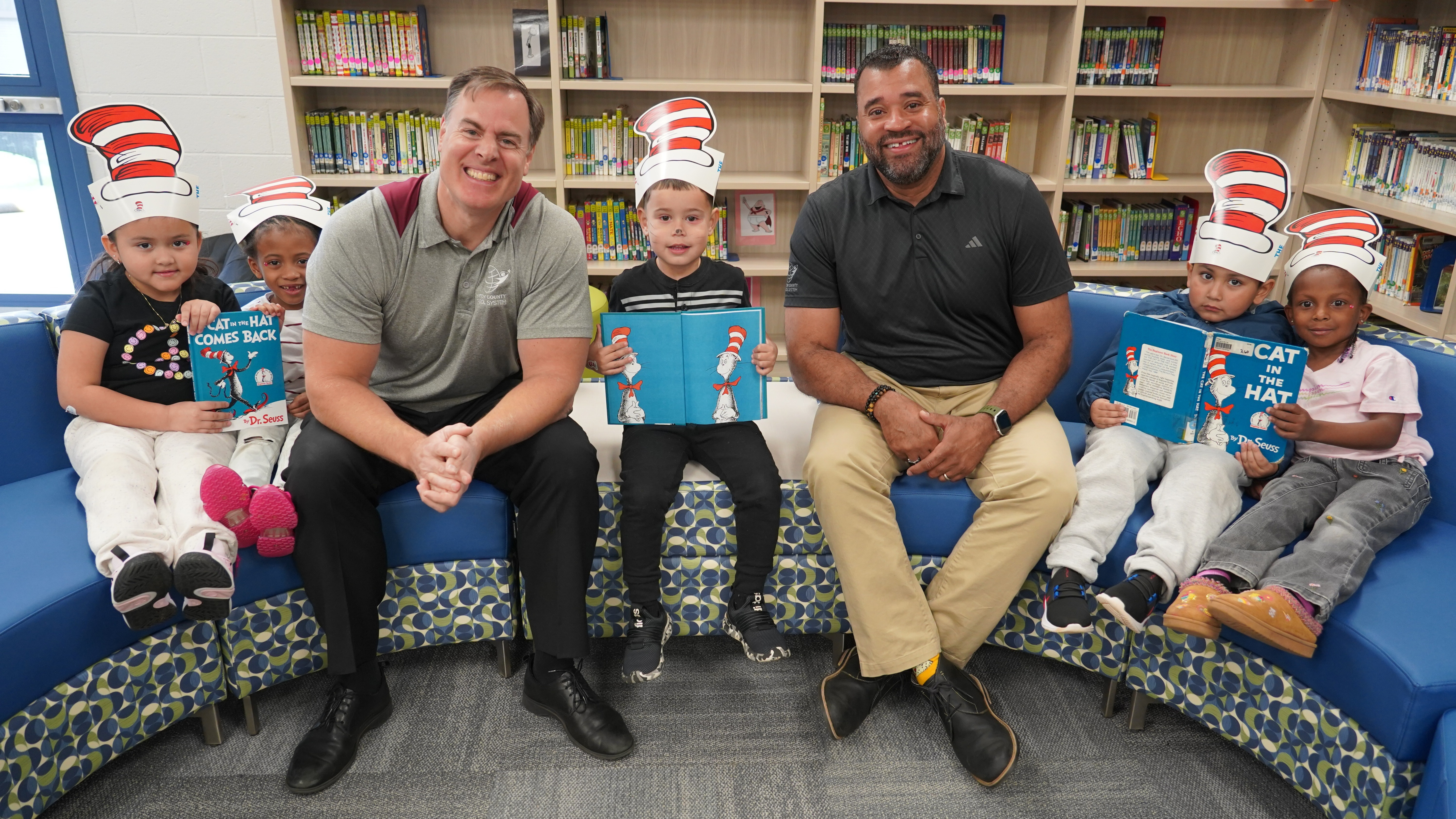 Men and children in Cat in the Hat hats sit on a blue bench in a library.