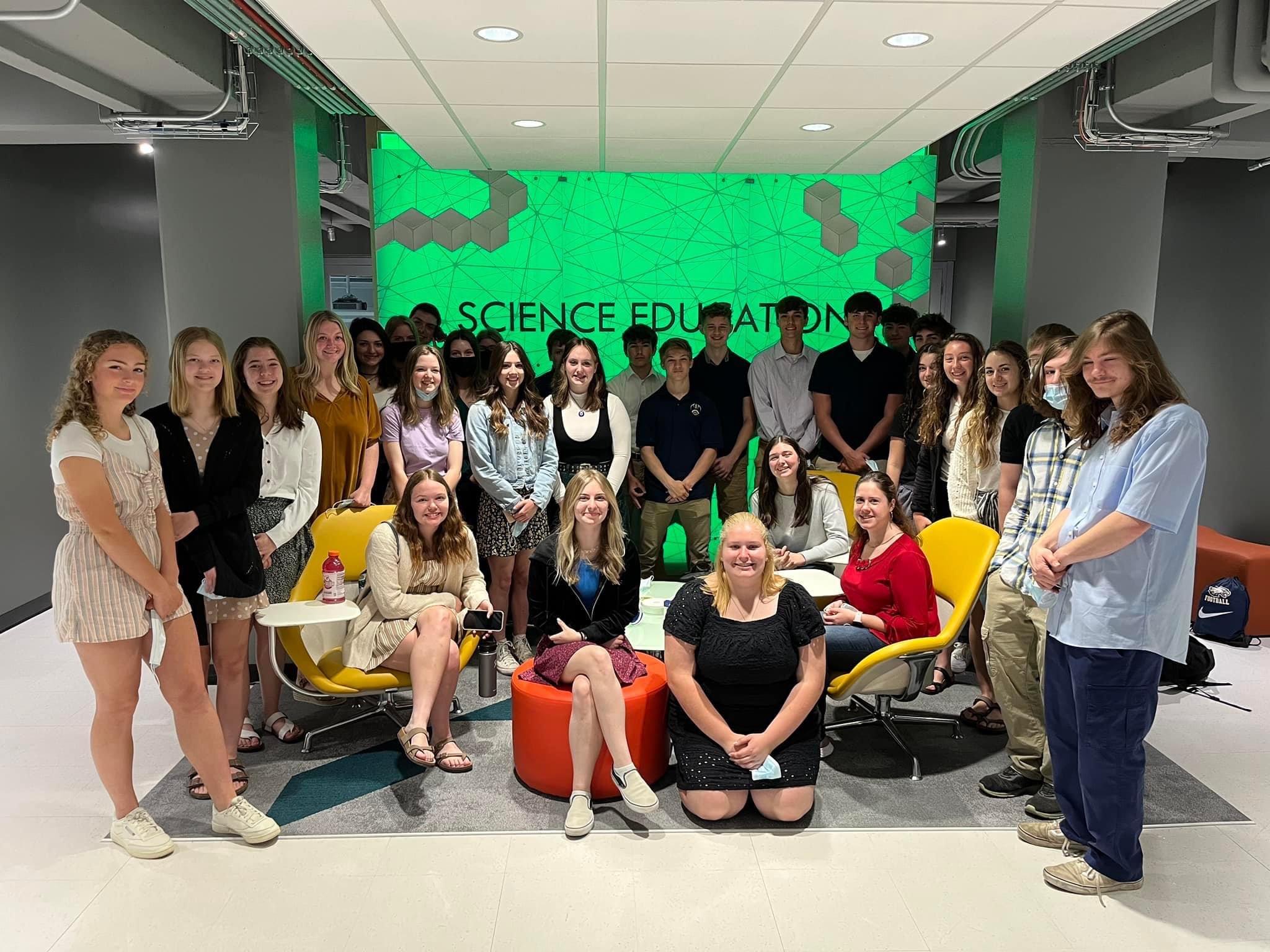 Students standing in a semi-circle around the center of the science education room. There is a neon green wall that lights up the background of the image, and the wall reads :science education".