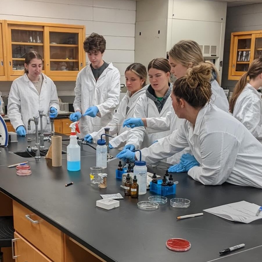 Lab students in white coats and gloves, some using microscopes, work at a lab bench with various equipment.