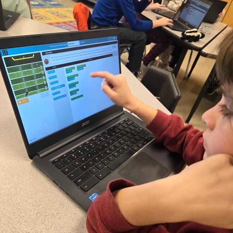 A child, seated at a table, points to a laptop screen displaying a graph. Behind, another person works at a table with multiple laptops.
