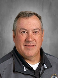 A man with gray hair wearing a gray and black jacket is smiling at the camera. He is standing against a gray background.