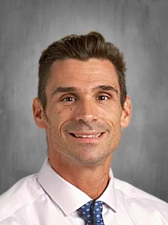 Man in a white shirt and blue tie with a light smile against a gray background.