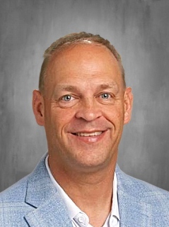 Man with short blond hair, smiling, wearing a light blue blazer and white shirt, standing in front of a gray background.