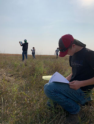 A group of individuals is engaged in an outdoor activity on a grassy field, with one person kneeling and writing in a notebook while others stand in the background holding materials.